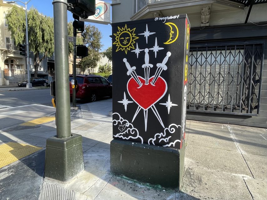 A utility box painted with a red heart pierced by five swords, surrounded by clouds, stars, a sun, and a crescent moon on a city street corner.