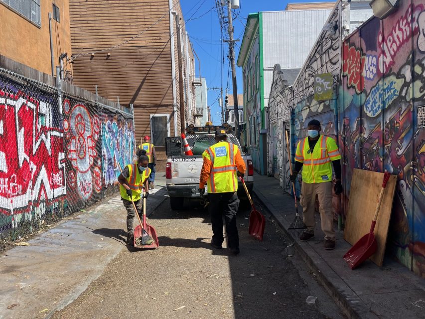 Four workers in safety vests clean a graffiti-covered alley near 16th and Mission with shovels, standing by a utility truck on a sunny day.