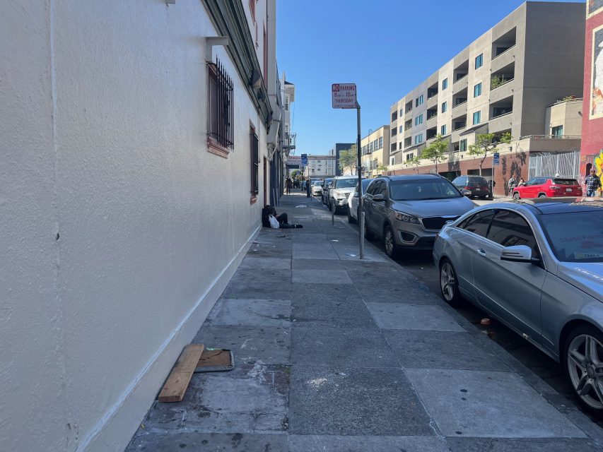 A city sidewalk with parked cars on the right and a person sitting on the ground near a building on the left; apartment buildings are visible in the background on a clear day.
