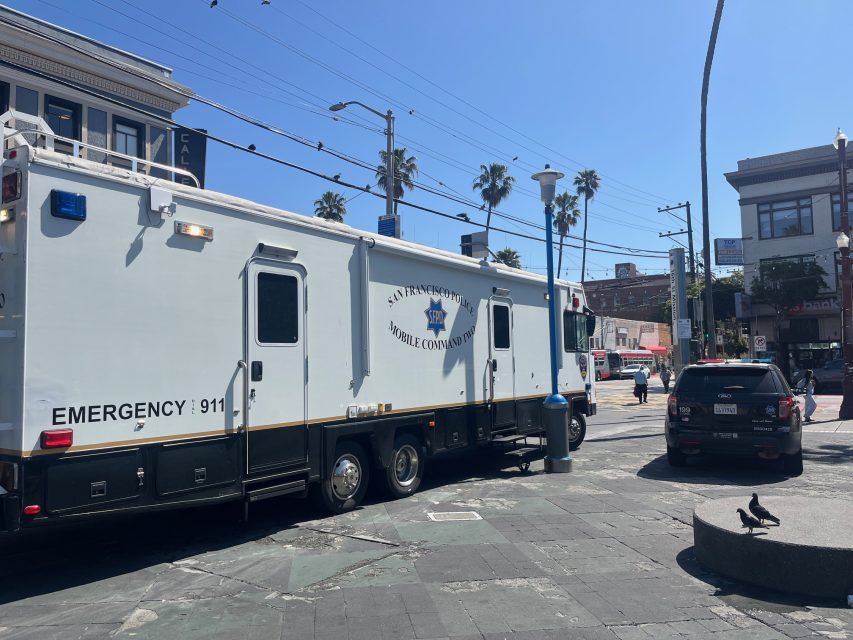 A San Francisco Police Mobile Command vehicle is parked at an intersection on a sunny day, with nearby buildings, palm trees, and pedestrians visible.