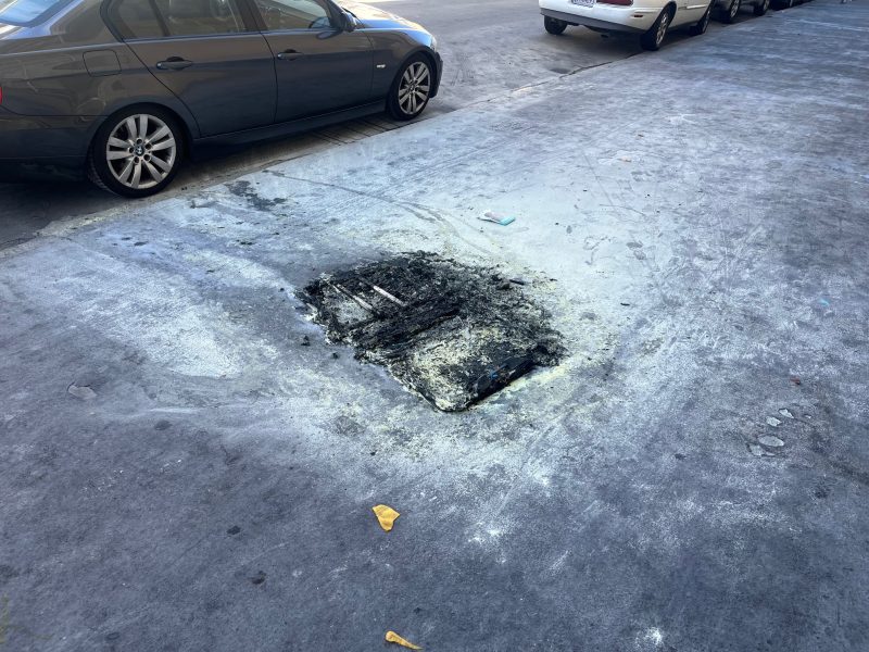 A large, burnt patch is visible on a city sidewalk near parked cars, with debris and scorch marks surrounding the area.