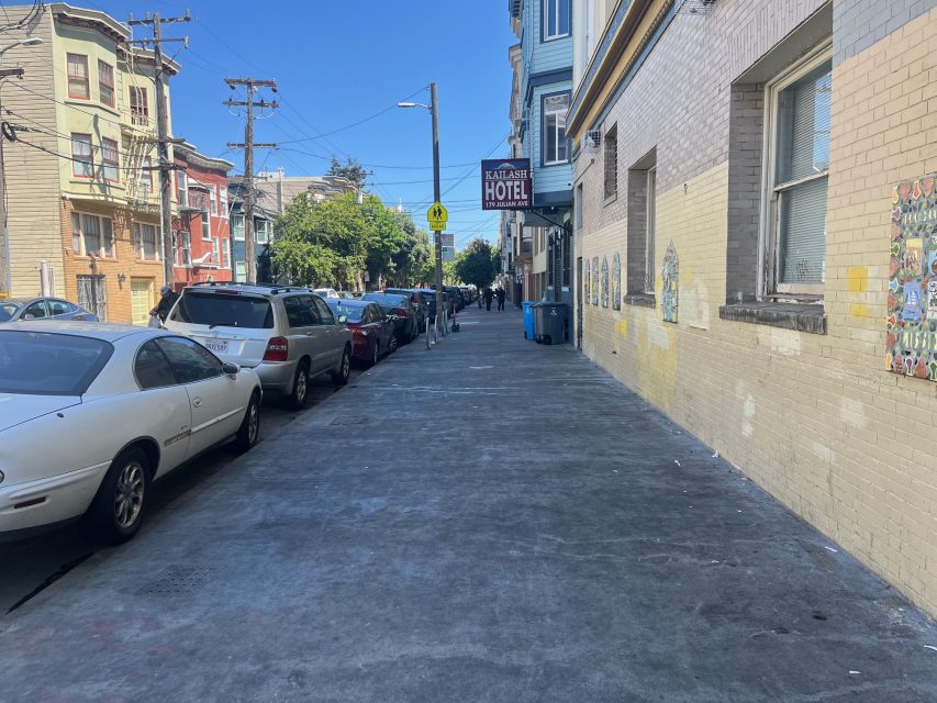 A city sidewalk lined with parked cars, apartment buildings, and a sign for the Kailash Hotel under a clear blue sky.