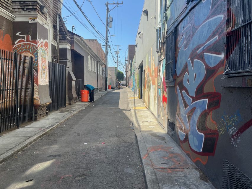 A narrow urban alley with graffiti-covered walls, gated doorways, a trash bin, overhead utility lines, and a clear blue sky.