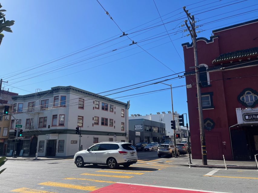 A white SUV waits at a crosswalk on a sunny day at an urban intersection with buildings, power lines, and clear blue sky.