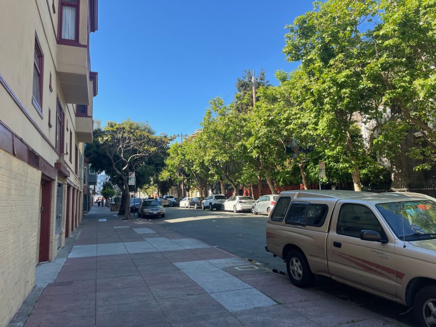 A sunny urban street with parked cars, a beige building on the left, and leafy trees lining the right side of the road.