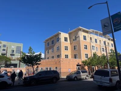 A multi-story beige and tan apartment building on a sunny day, with parked cars along the street and several people gathered near the entrance.