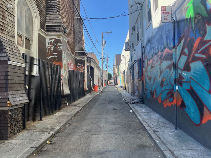 Narrow urban alley with graffiti-covered walls, a "No Parking Any Time" sign, overhead wires, trash bins, and some debris scattered on the ground under bright daylight.