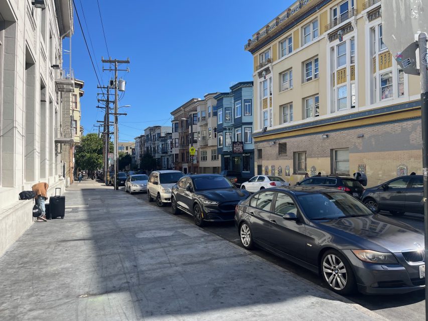 Cars parked along a sunny city street lined with multi-story buildings; a person stands with bags near the sidewalk on the left.