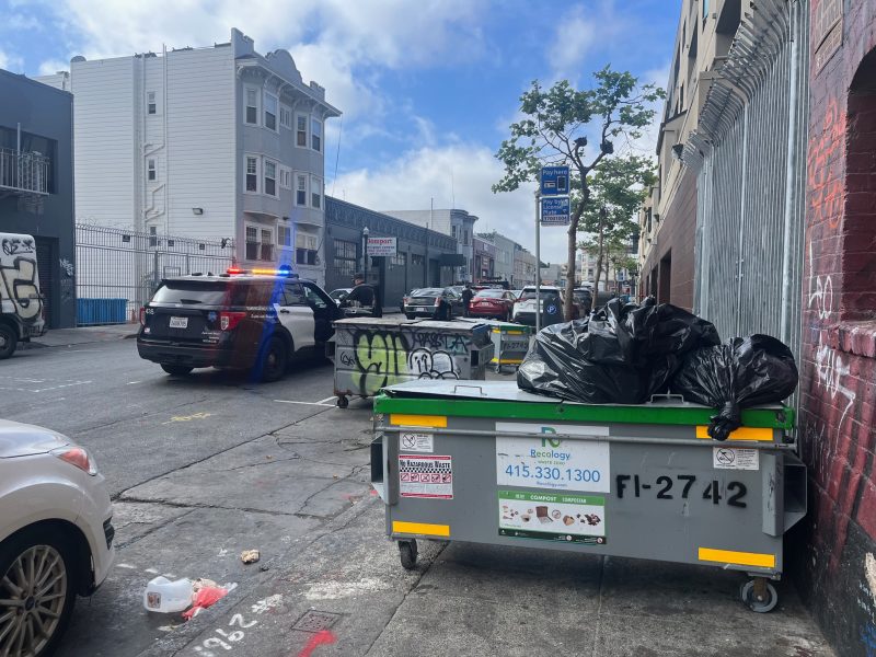 A police car with flashing lights is parked on a city street near a green dumpster filled with trash bags, with graffiti visible on nearby walls and buildings.