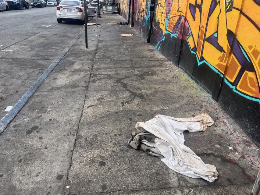 A dirty sheet lies on a litter-strewn sidewalk next to a graffiti-covered wall, with parked cars visible further down the street.