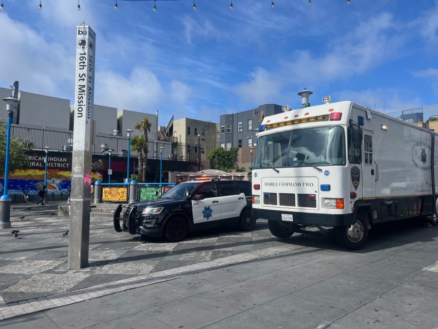 A San Francisco police car and a mobile command vehicle are parked at 16th St Mission plaza, with nearby buildings and murals visible in the background.