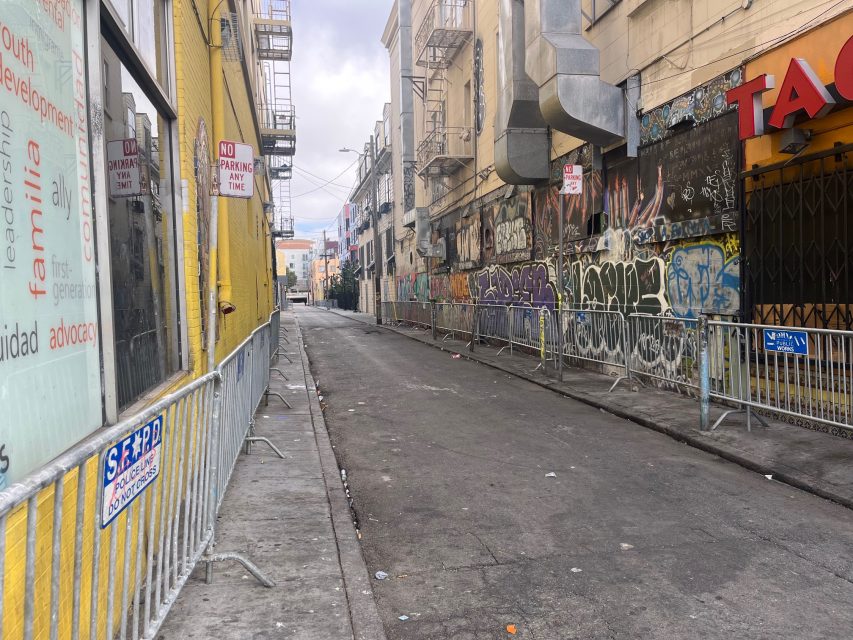 Narrow urban alleyway with graffiti-covered walls, metal barricades, and various signs, including "No Parking Any Time." The street is mostly empty and lined with buildings.