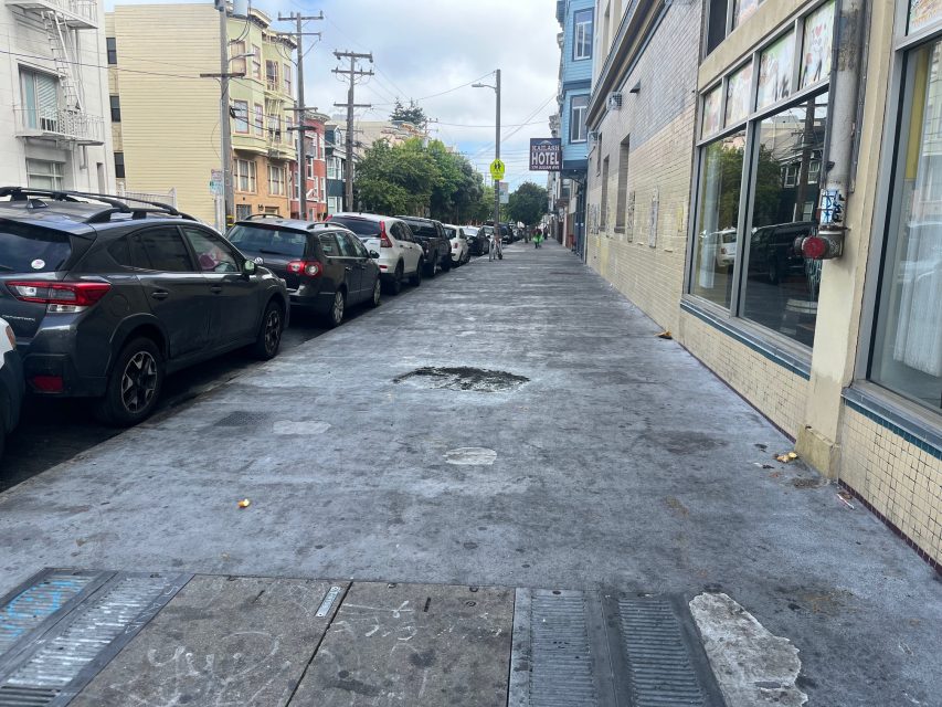 A city sidewalk with a large hole or damaged area in the pavement, lined with parked cars and buildings on both sides.