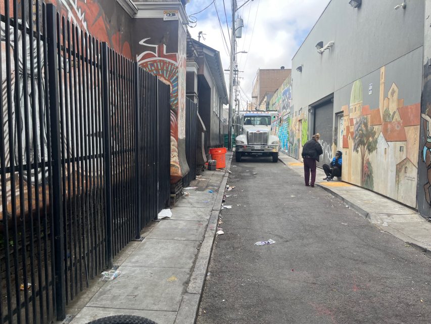 A narrow urban alley with murals on both sides, scattered trash, a garbage truck, and a few people walking or sitting along the sidewalk.
