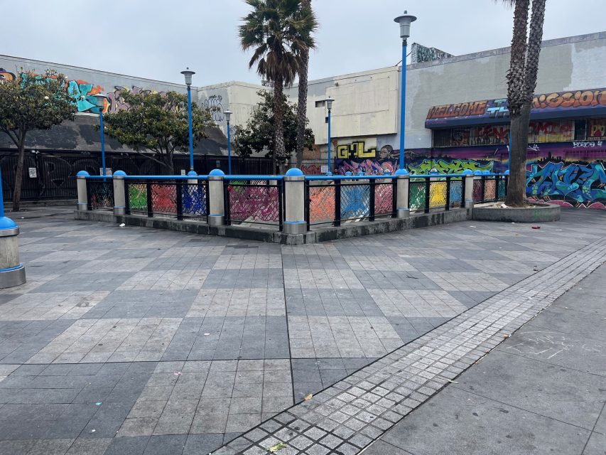 Urban plaza with gray tiled pavement, blue railings featuring rainbow panels, palm trees, and buildings covered in colorful graffiti.