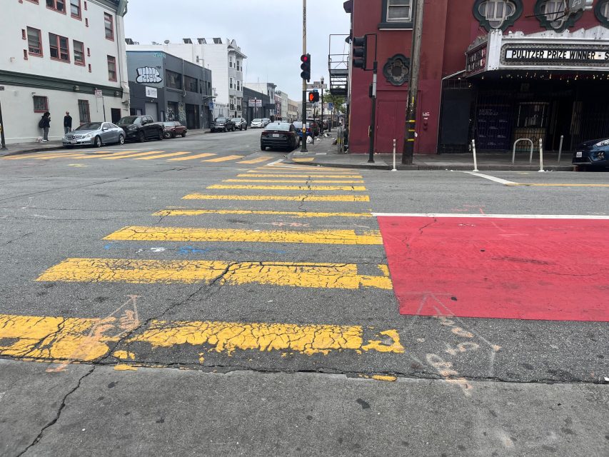 A crosswalk with faded yellow stripes and a red painted curb at a city intersection; buildings and parked cars are visible in the background.