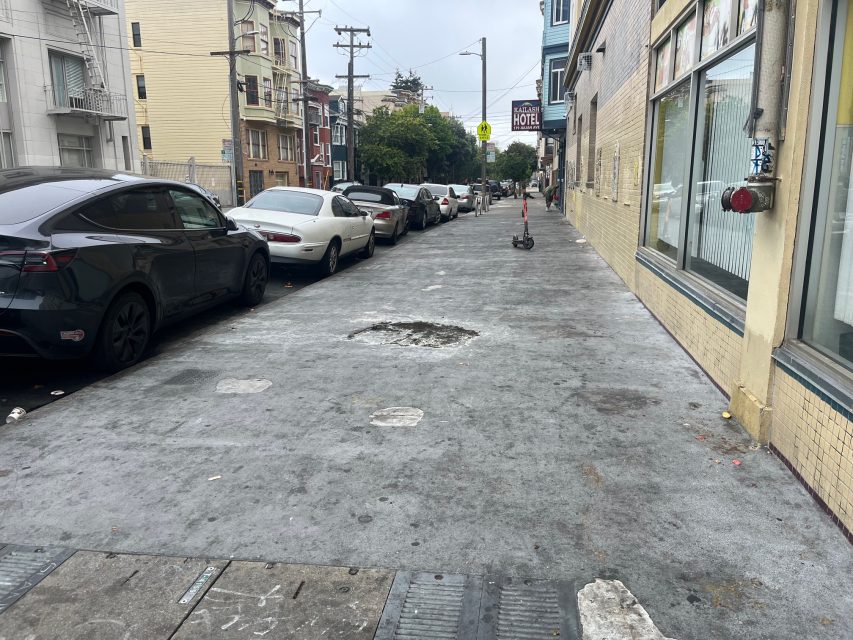A city sidewalk with a damaged patch in the center, lined with parked cars on one side and buildings on the other; a scooter is near the background.