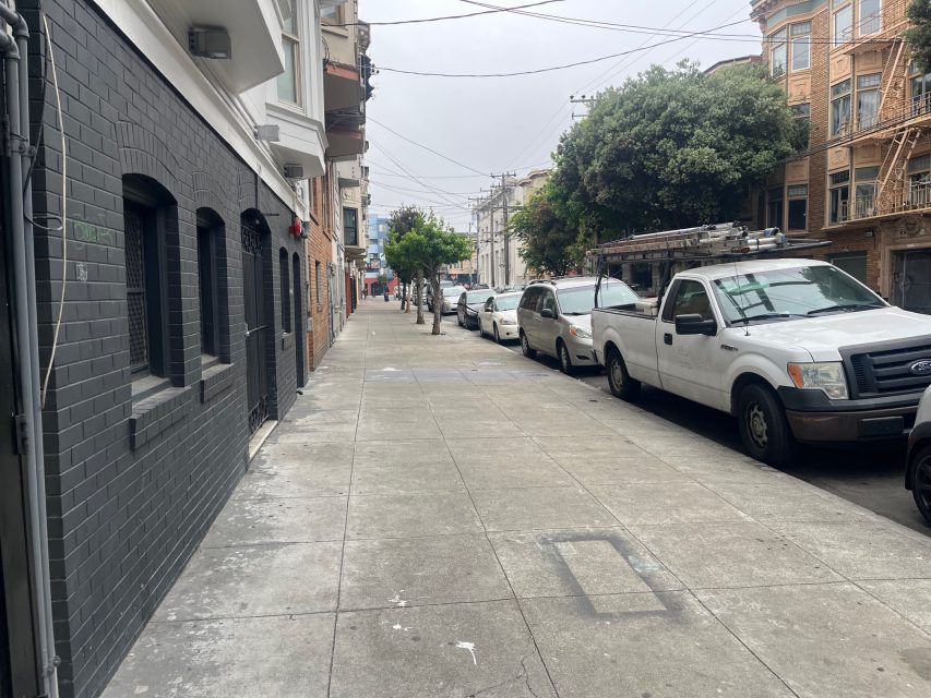 A city sidewalk next to a black brick building, with several parked cars and trees lining the street on a cloudy day.