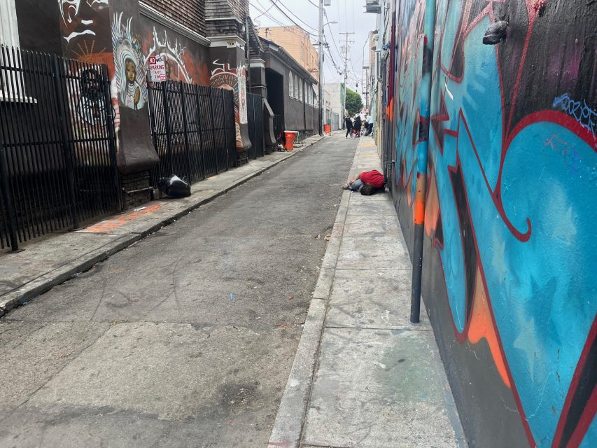 Narrow urban alley with graffiti on walls, litter on the ground, a person lying on the sidewalk, and several people visible in the distance.