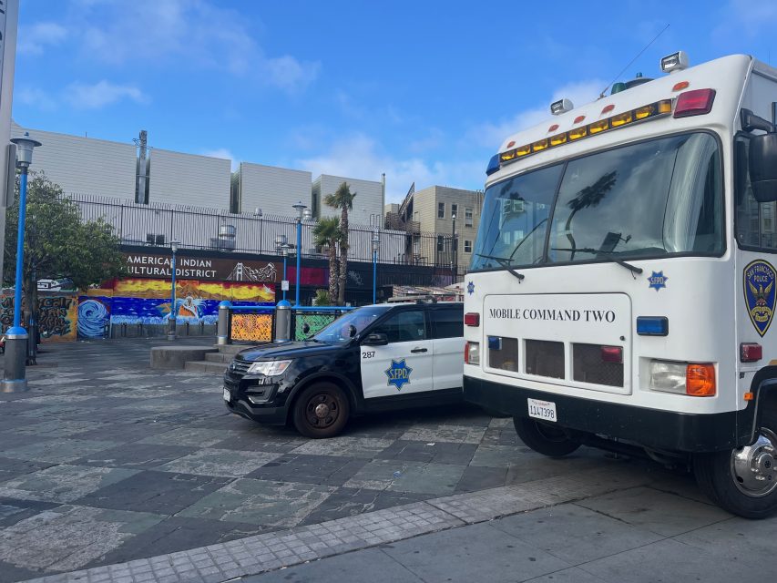 A police SUV and a mobile command vehicle are parked in a city plaza near a mural labeled "American Indian Cultural District.