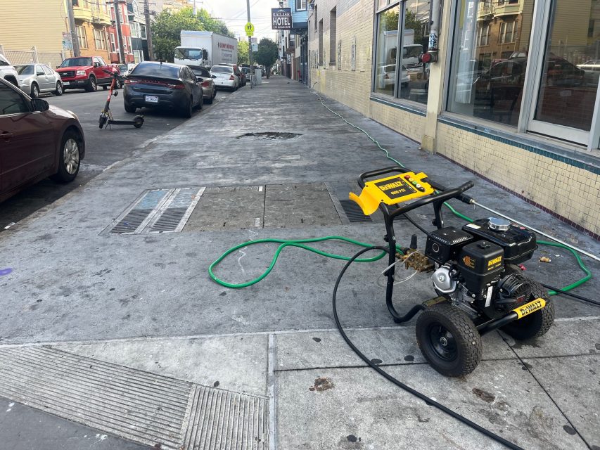 A pressure washer with hoses is set up on a city sidewalk next to a building; cars and a scooter are also visible along the street.