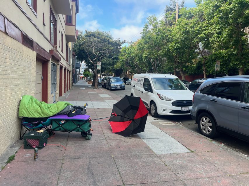 A green sleeping bag on a cot, a red and black umbrella, and personal items are set up on a city sidewalk beside parked cars and trees.