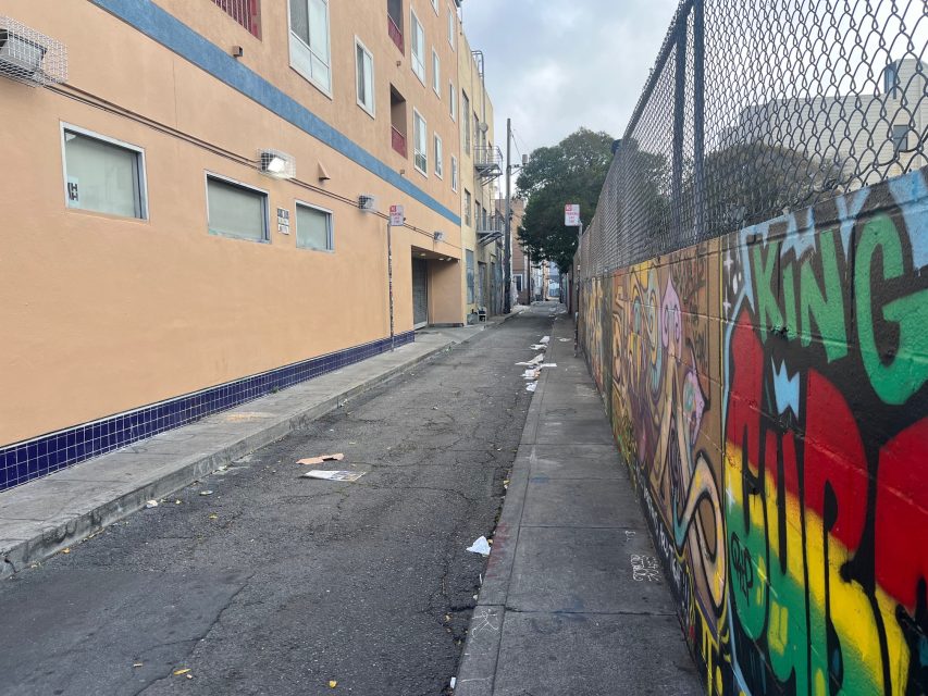 A narrow urban alleyway with scattered litter, a beige building on the left, and a fence with colorful graffiti on the right.