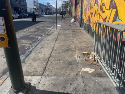 A city sidewalk with scattered litter, a bike rack, graffiti on the wall, and empty street in the background under daylight.