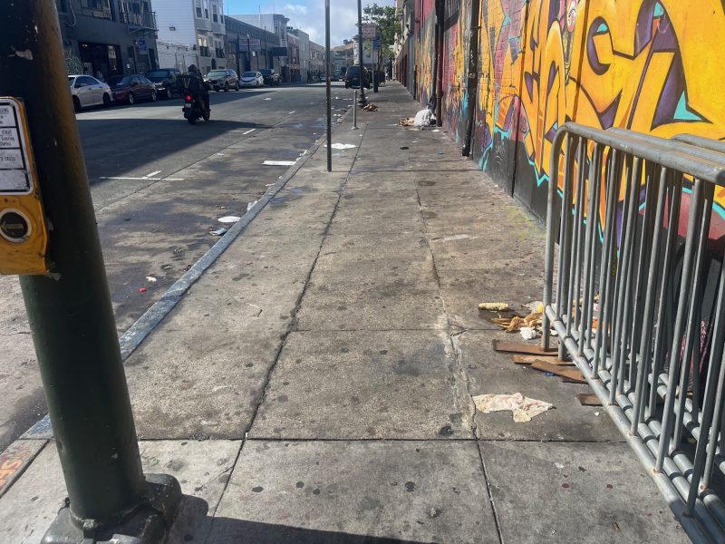A city sidewalk with scattered litter, a bike rack, graffiti on the wall, and empty street in the background under daylight.