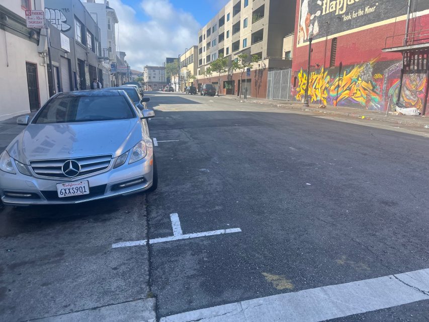 A silver Mercedes is parked on a city street near 16th and Mission, with graffiti on the wall to the right and several other cars ahead. The street appears mostly empty under a partly cloudy sky.