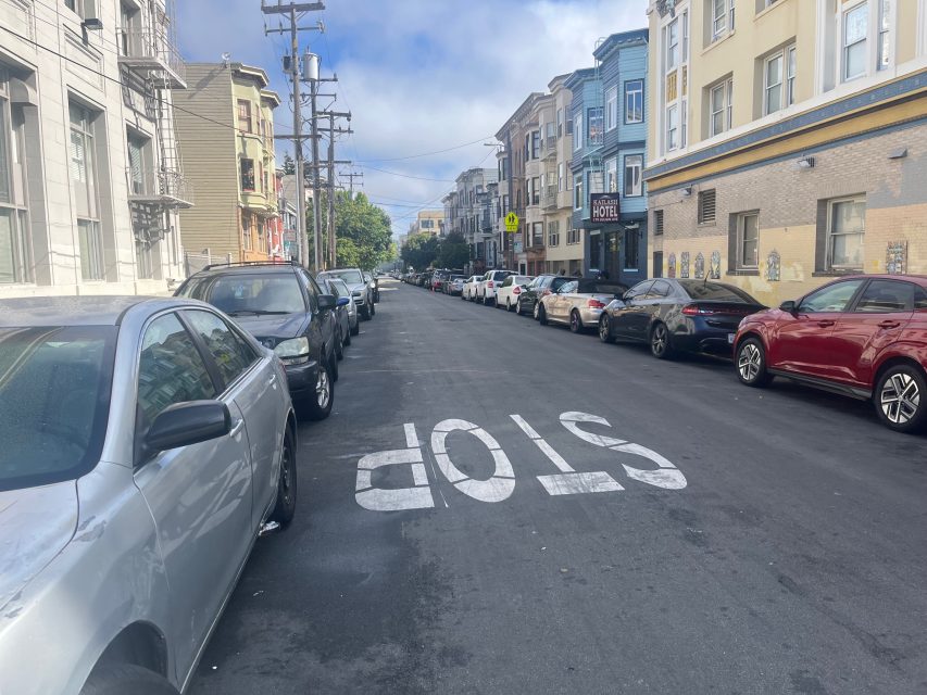 A city street with cars parked on both sides, apartment buildings lining the road, and the word "STOP" painted on the pavement in white.