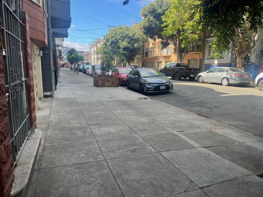 A quiet city street with parked cars on the right side, trees along the sidewalk, and buildings lining both sides of the street under a clear sky.