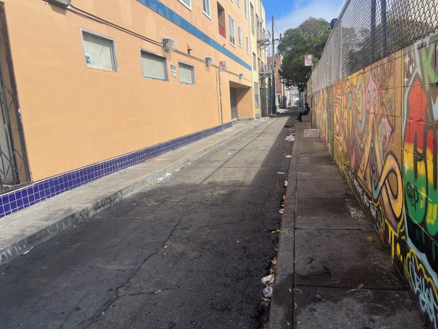 Urban alleyway with scattered litter, bordered by a building with barred windows on the left and a graffiti-covered wall on the right.