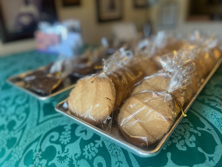 Two trays of cookies wrapped in clear plastic are displayed on a turquoise patterned tablecloth, with a blurred indoor background.