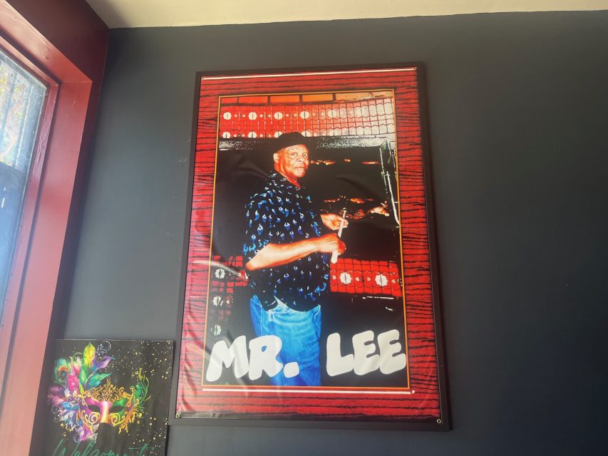 Framed photo on a dark wall showing a smiling man in a hat holding drumsticks, with the name "MR. LEE" in large white letters at the bottom.