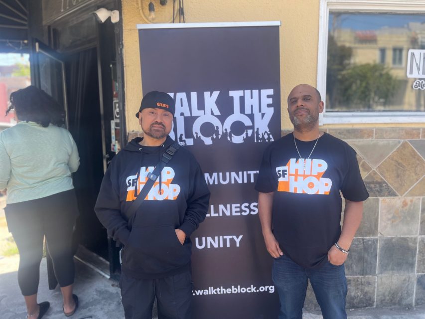 Two men stand in front of a "Walk the Block" banner; both wear black "SF Hip Hop" shirts and face the camera outside a building.