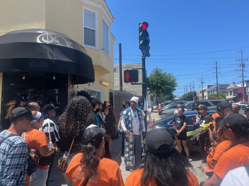 A group of people gathers on a street corner near a café to listen to a speaker in traditional attire under a traffic light on a sunny day.