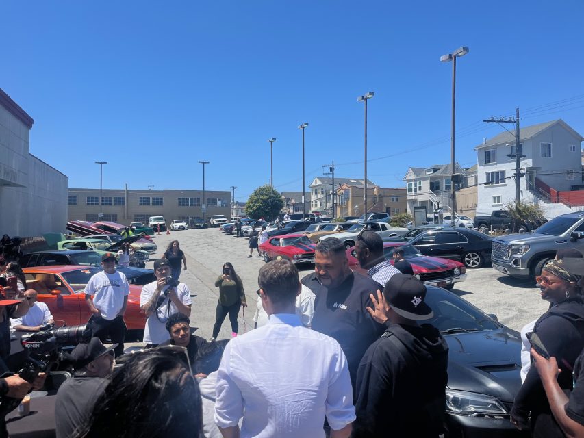 A group of people gathers around several parked cars in a sunny outdoor lot, with more cars and buildings visible in the background.