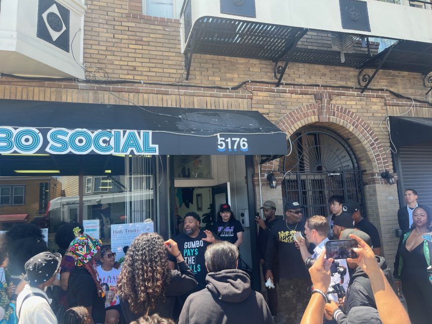 A crowd gathers outside a storefront with the sign "GUMBO SOCIAL" at 5176; people are talking, taking photos, and holding signs in daylight.