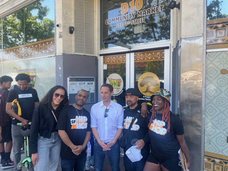 A group of people pose in front of the D10 Community Market entrance; some wear matching "SF Hip Hop" shirts.