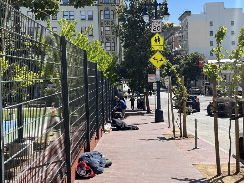 Several people are lying or sitting on a city sidewalk next to a fenced area, with bags and belongings nearby; school zone signs and cars are visible along the street.