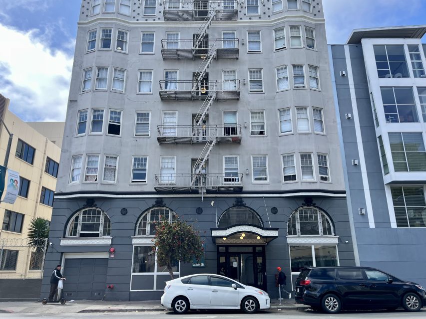 A multi-story gray apartment building with arched windows on the ground floor, fire escapes, and cars parked along the street in front.