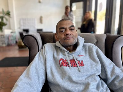 A man in a gray Ole Miss hoodie sits on a brown armchair in a well-lit room with people and plants in the background.