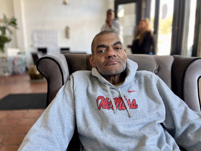 A man in a gray Ole Miss hoodie sits on a brown armchair in a well-lit room with people and plants in the background.