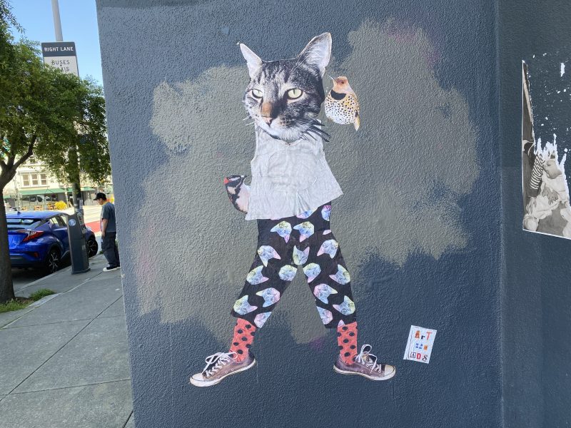 Street art of a cat with a bird on its shoulder, wearing patterned pants, red polka-dot socks, and sneakers, pasted on a gray painted wall at a street corner.