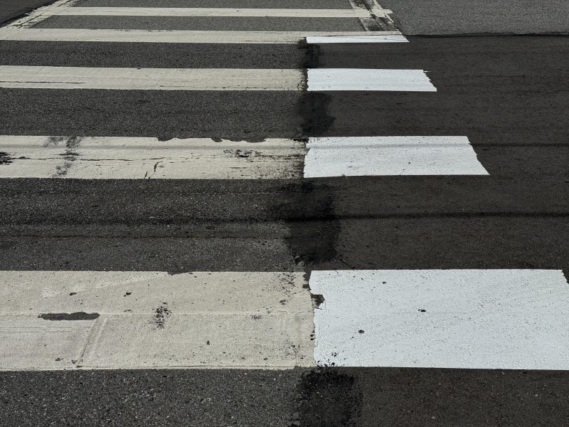 A crosswalk with faded and newly repainted white stripes crosses an asphalt road, showing a contrast between old and fresh paint.