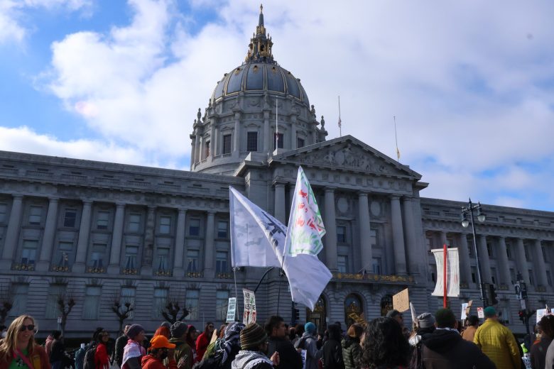 A crowd of people with flags and signs gathers in front of a large government building with a prominent dome under a partly cloudy sky.