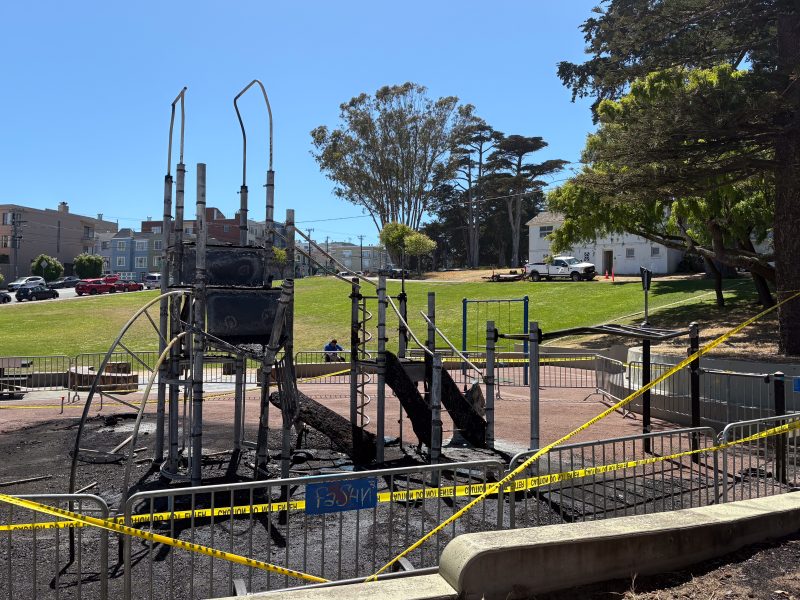 Charred remains of a playground structure after a playground fire, surrounded by caution tape in a park, with grass and trees in the background.