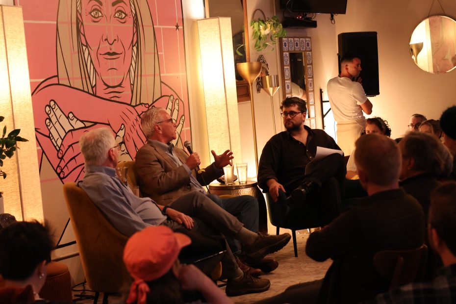 Three men sit and talk in front of an audience at a panel discussion, with a large illustrated mural behind them and warm lighting in the room.