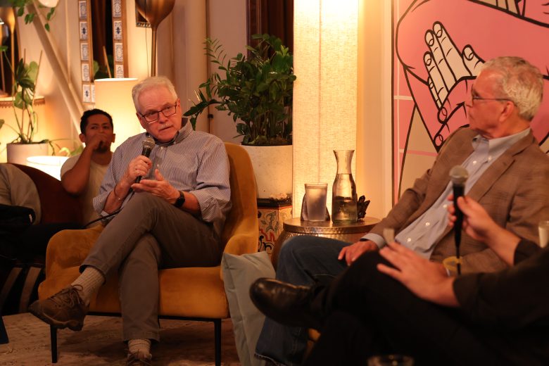 Two men are seated, speaking into microphones during a panel discussion in a cozy, well-lit room with plants and artwork in the background.